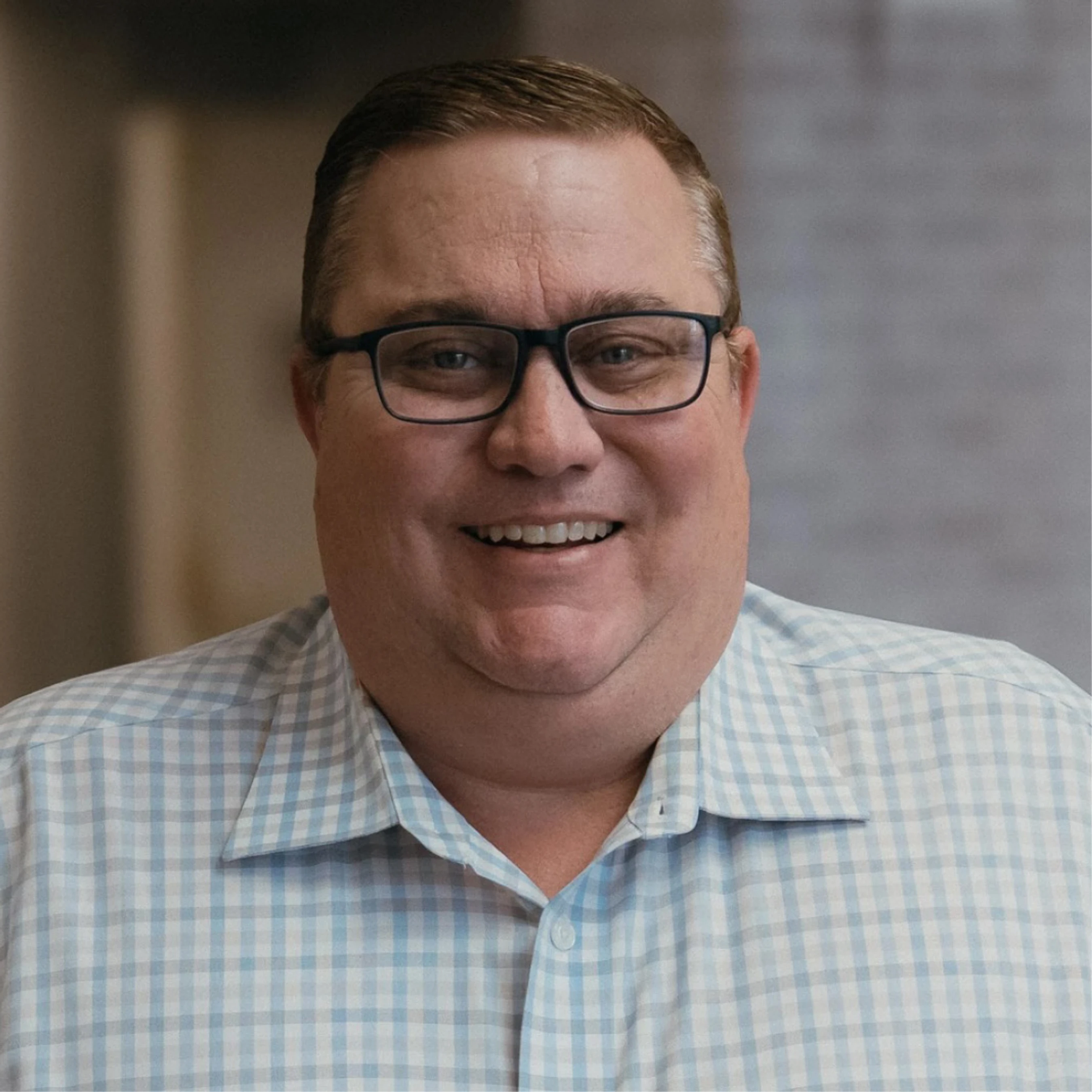 A close-up portrait of a middle-aged white man with short brown hair and glasses, smiling at the camera while wearing a blue and white checkered shirt.