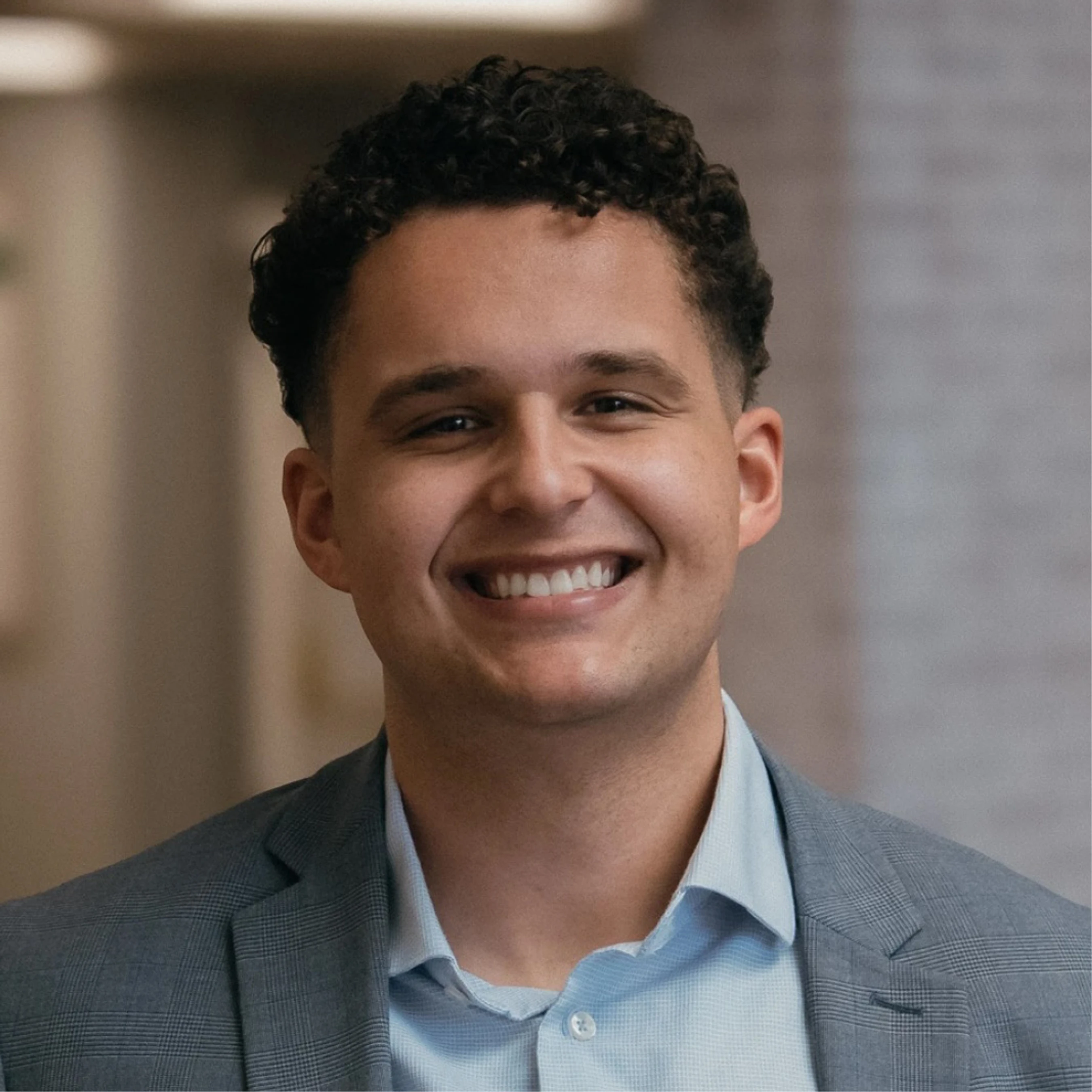 A headshot of a young man with dark curly hair smiling warmly at the camera. He is wearing a light blue button-down shirt and a grey plaid blazer.