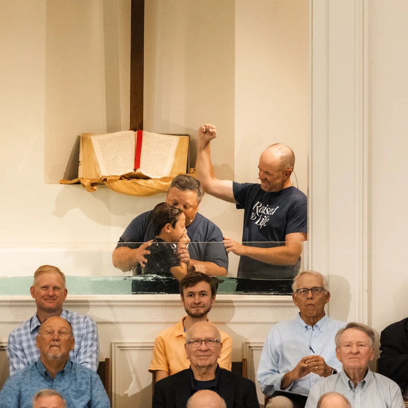 A young boy is held by a man in a baptismal font while another man raises his fist in celebration. A congregation of men watches from the pews.