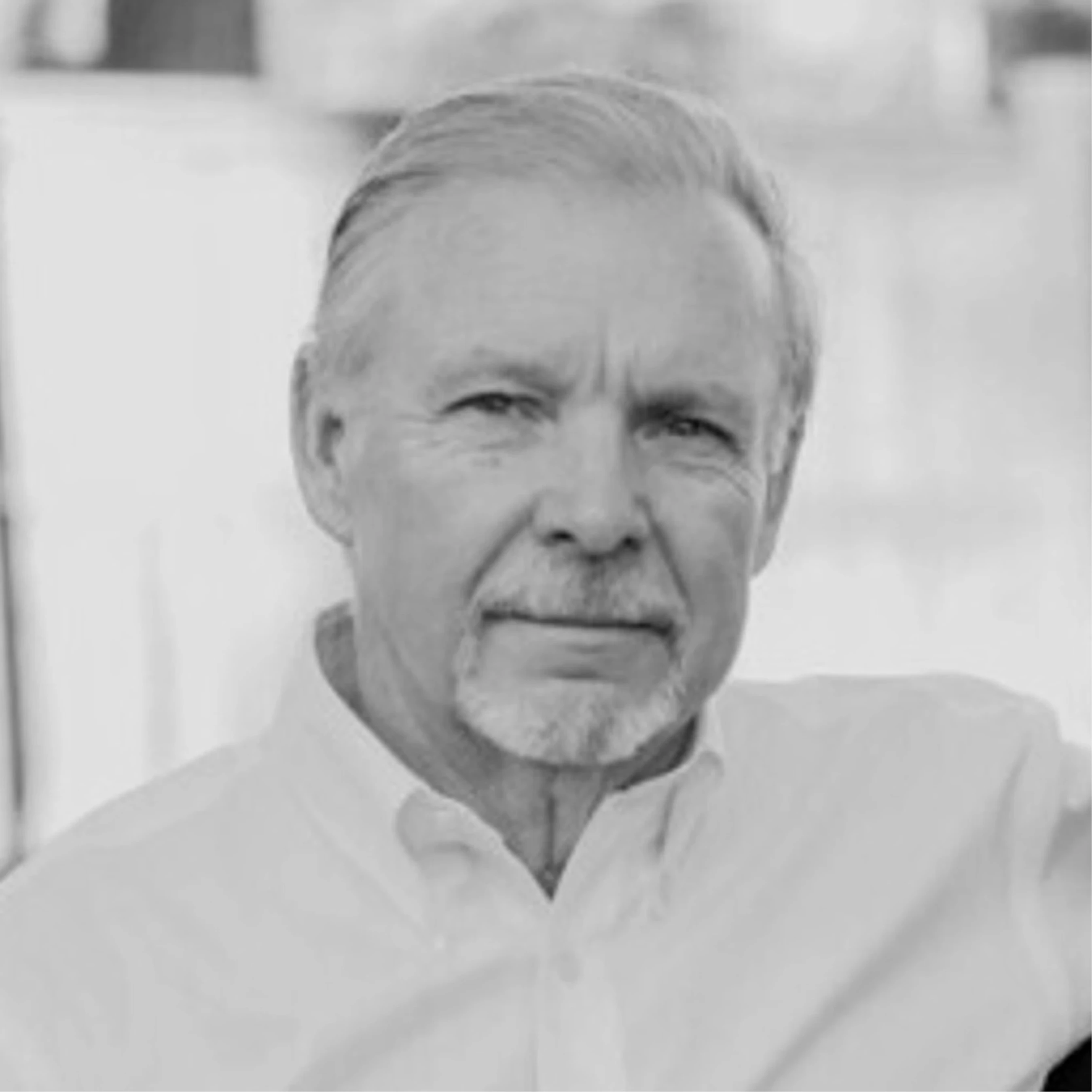 A black and white headshot of an older man with graying hair, a mustache, and a goatee. He is wearing a white collared shirt and looking at the camera.