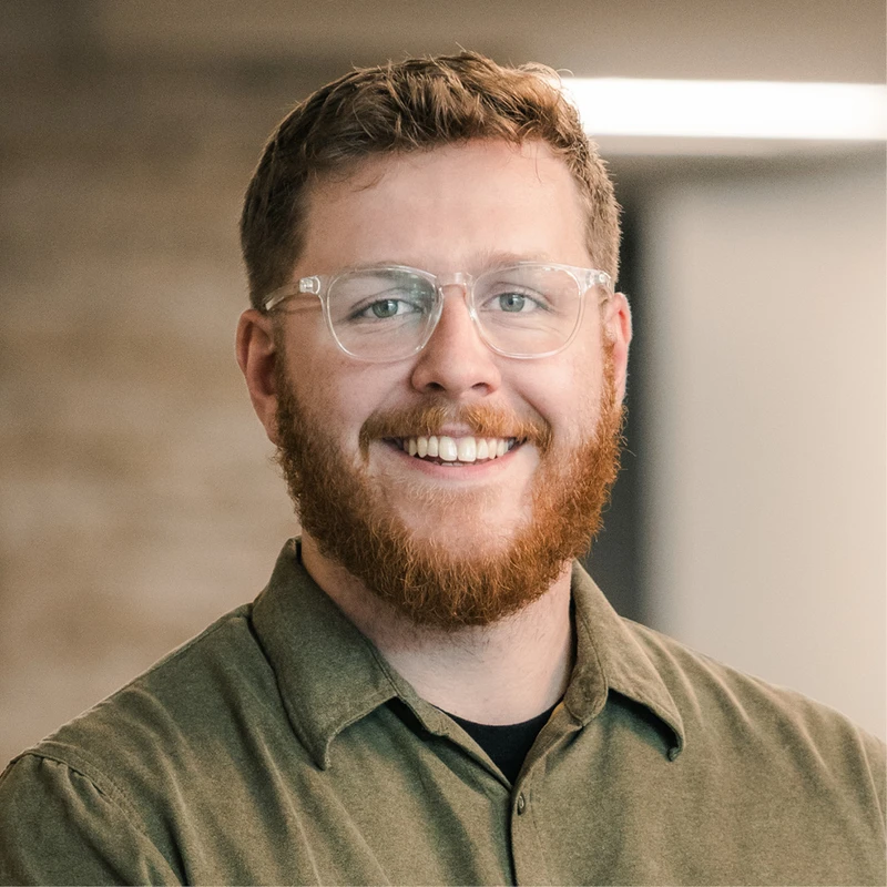 A headshot of a friendly-looking man with red hair, a full red beard, and clear-framed glasses. He is smiling at the camera and wearing an olive green shirt.