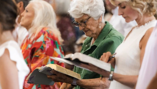 An elderly woman with white hair and glasses focuses on reading her open Bible while standing in a church congregation during a service.