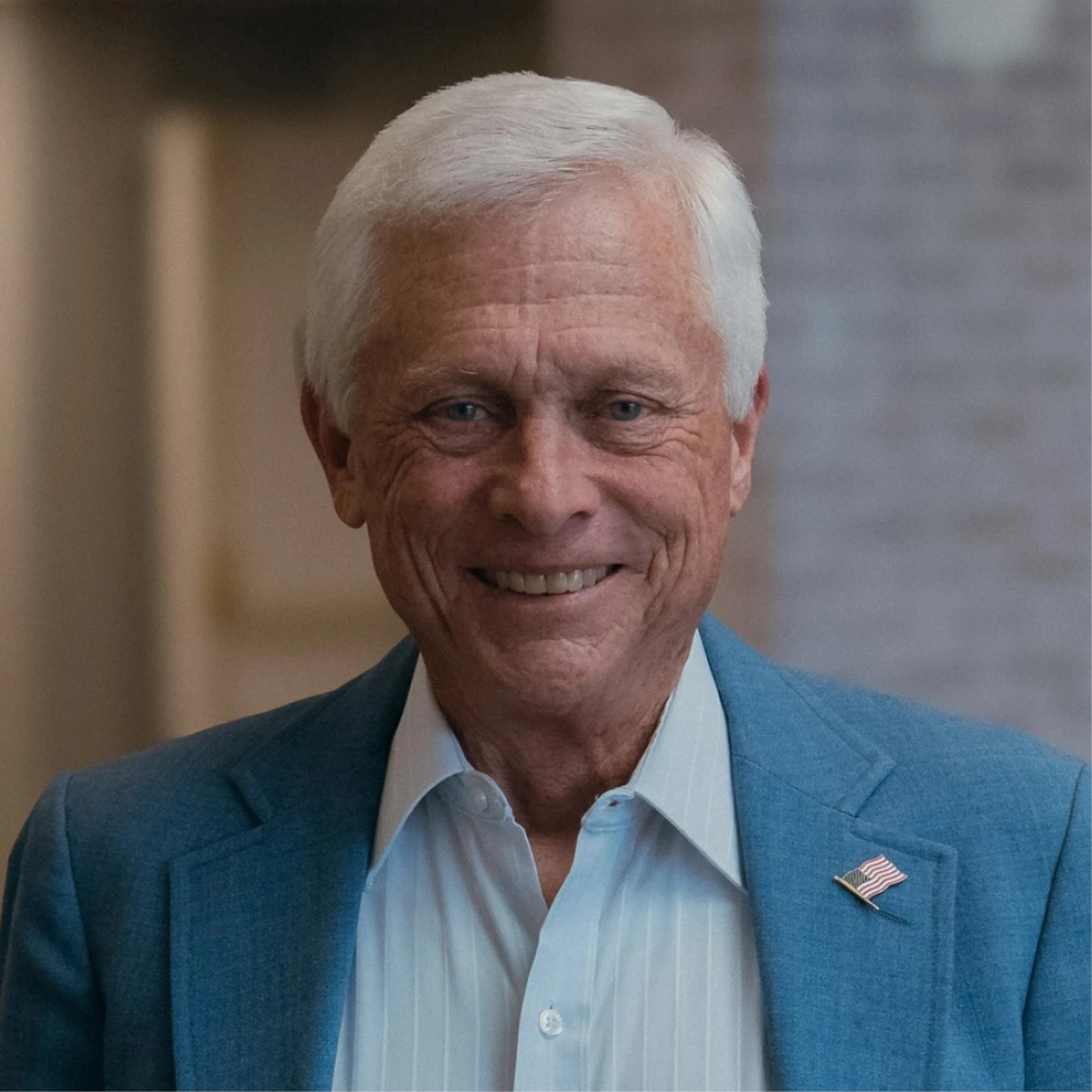 A headshot of an older man with white hair smiling warmly. He is wearing a blue blazer with an American flag pin over a light blue and white striped shirt.