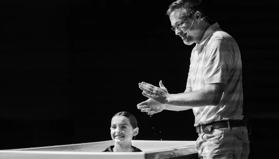 A black and white photo of a smiling young girl in a baptismal pool, looking up at a pastor who is smiling and clapping his hands beside her.