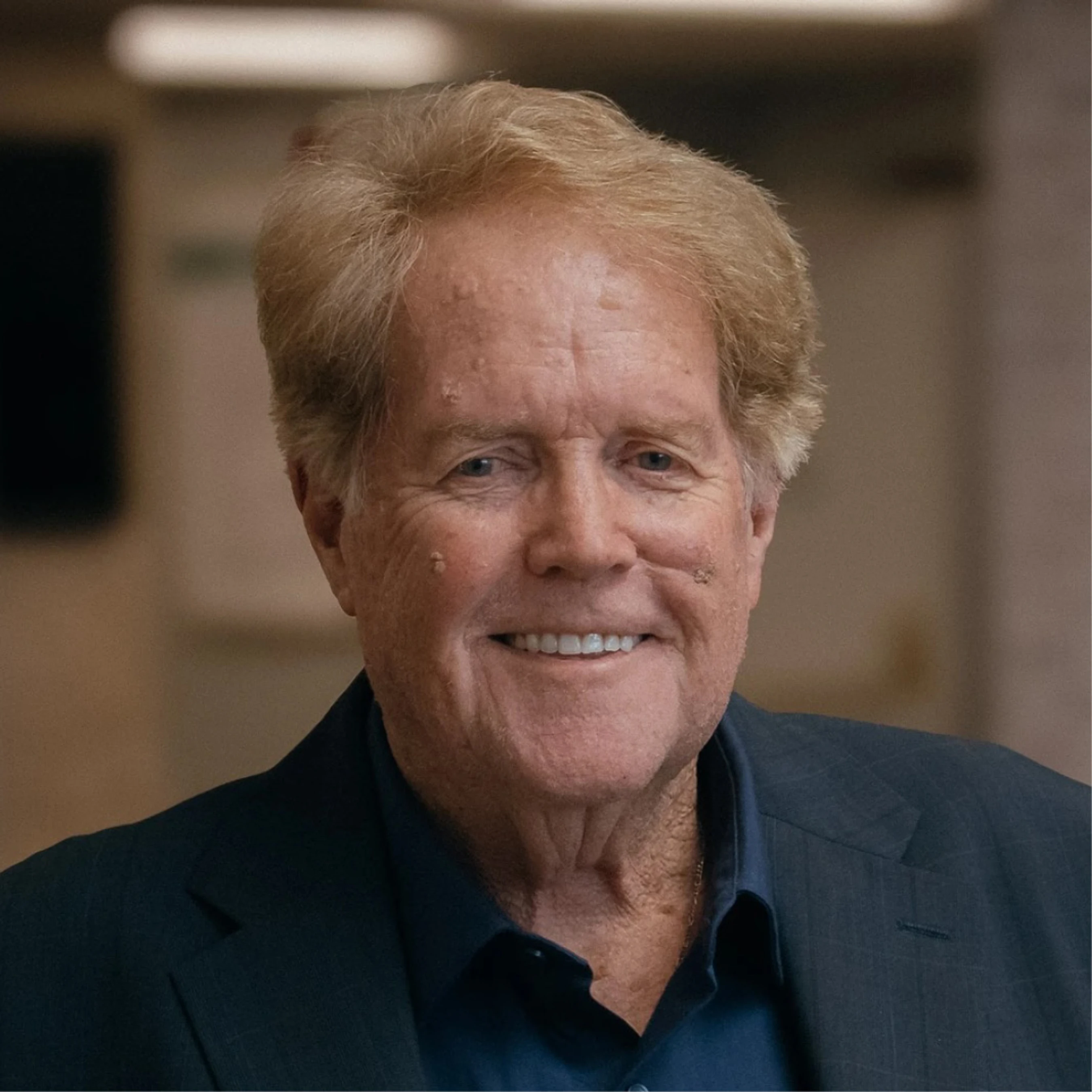 A headshot of an older man with blonde, styled hair, smiling warmly. He is wearing a dark blue suit jacket over a dark blue collared shirt.