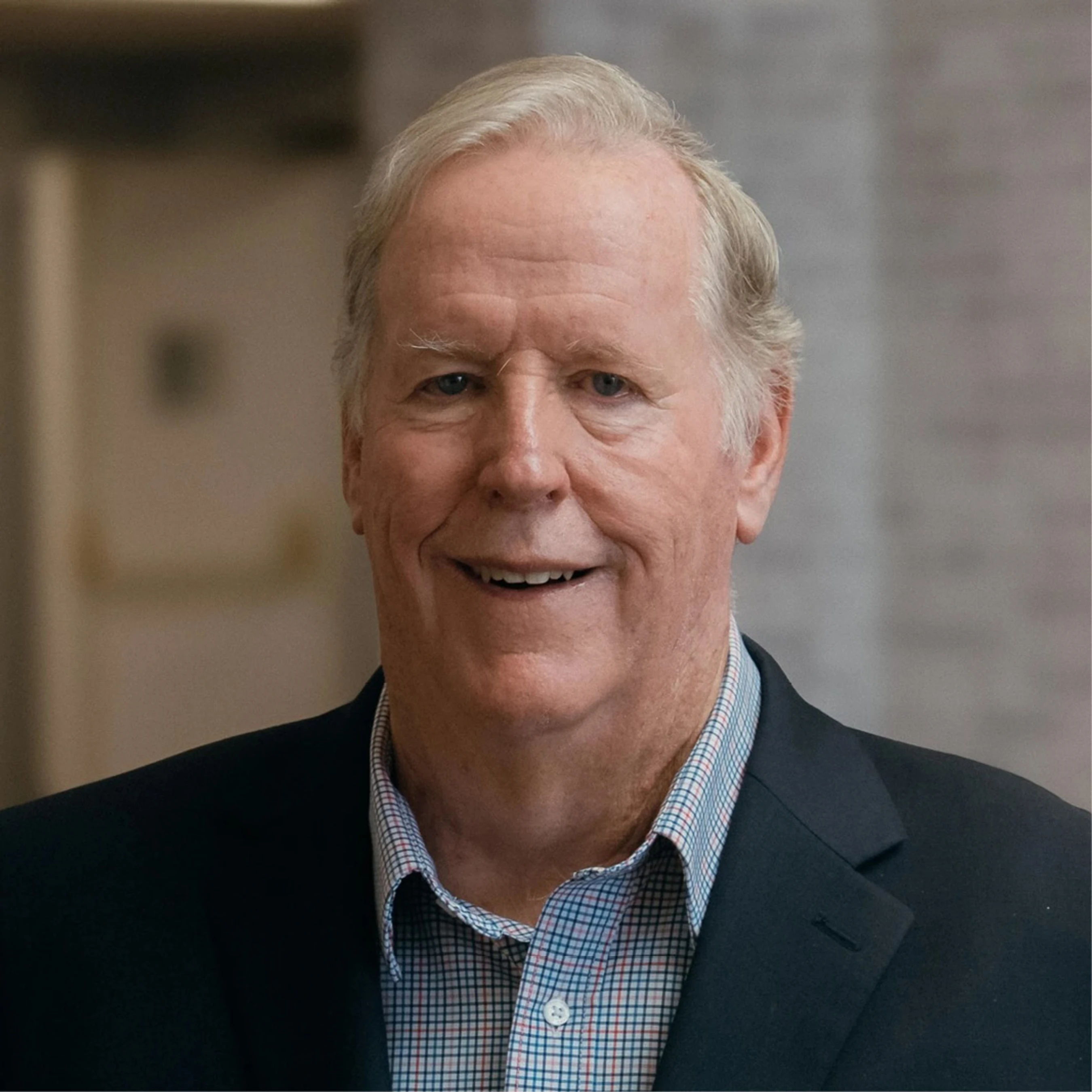 A head and shoulders portrait of an older white man with grey hair, smiling at the camera. He wears a dark blazer over a checkered shirt.