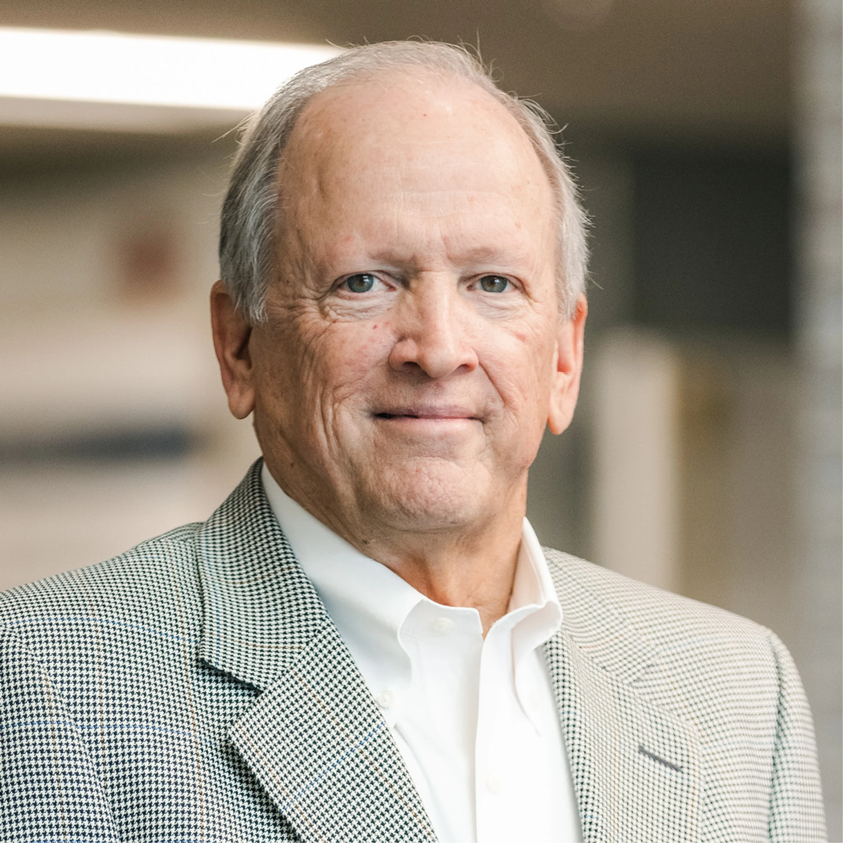 A head-and-shoulders portrait of an older man with gray hair, smiling slightly. He is wearing a white collared shirt under a houndstooth blazer.