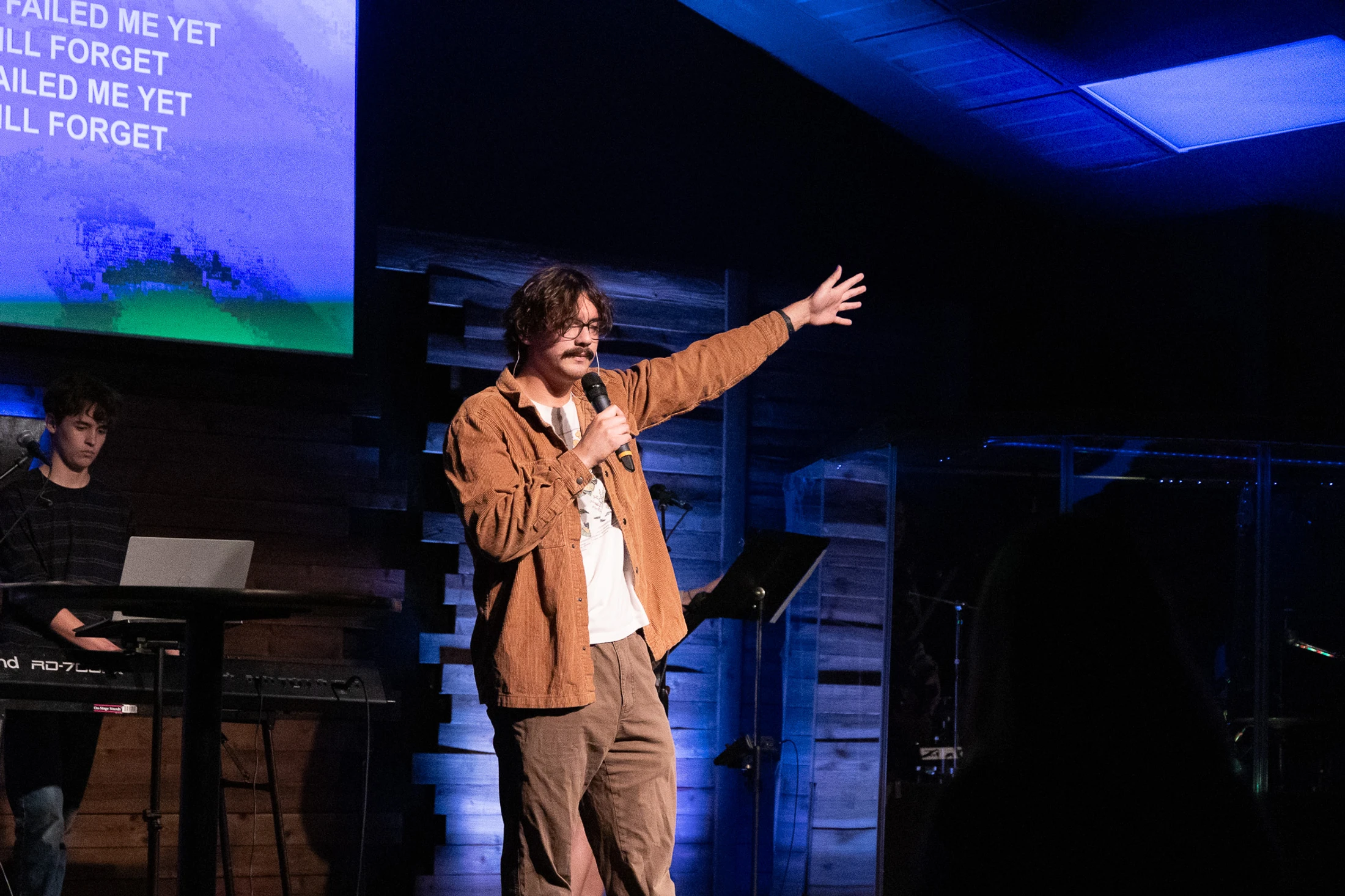 A man with a mustache and glasses in a brown jacket speaks into a microphone on stage, gesturing with his arm. A screen with lyrics is behind him.