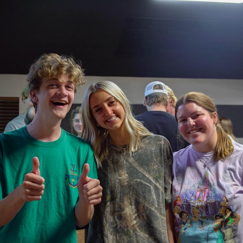 Three young people stand together and smile at the camera. The person on the left wears a green shirt and gives two thumbs up.