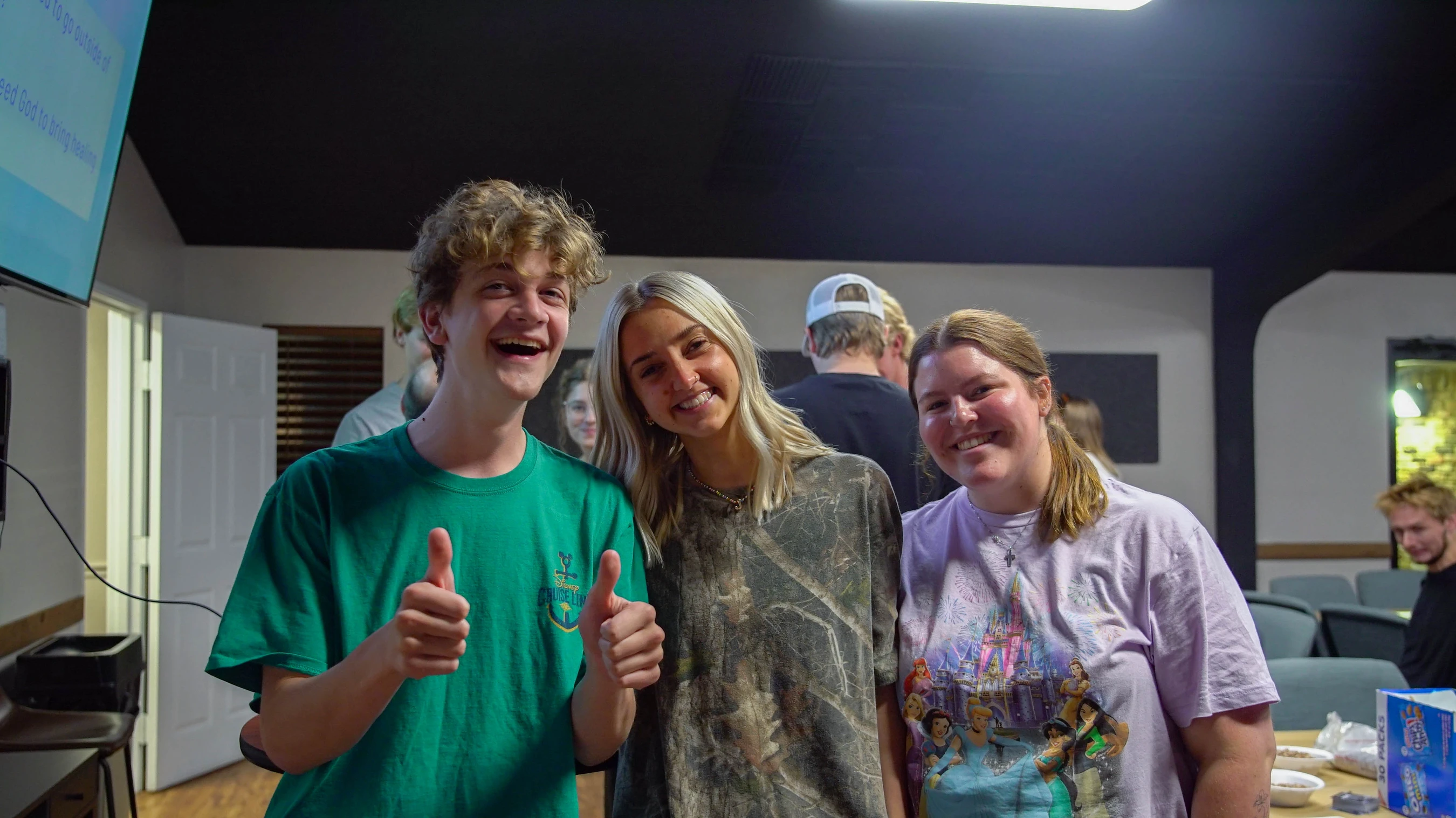 Three young people stand together and smile at the camera. The person on the left wears a green shirt and gives two thumbs up.