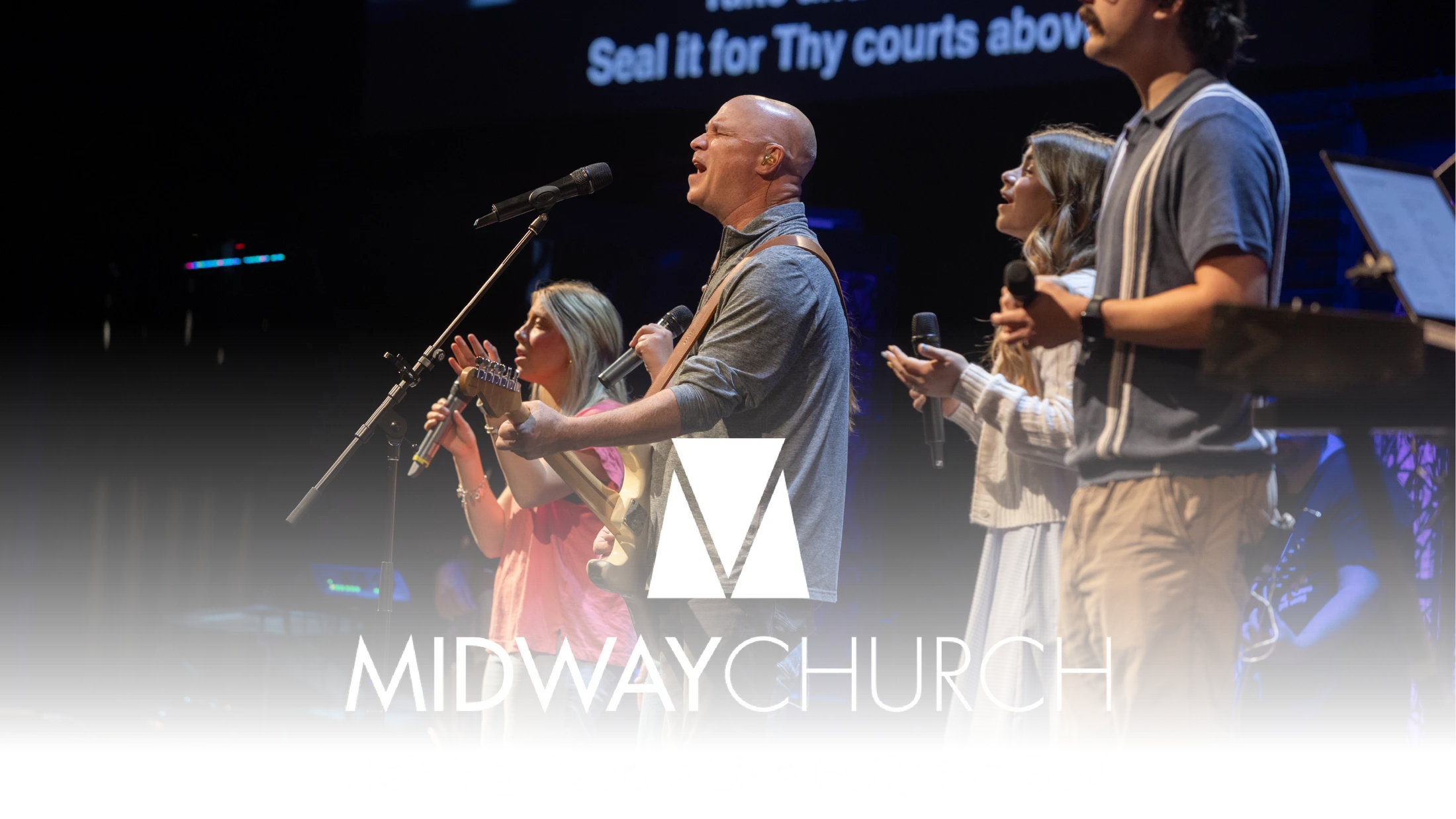A worship band performs on stage at Midway Church. A bald man sings passionately into a microphone, surrounded by other vocalists and musicians.