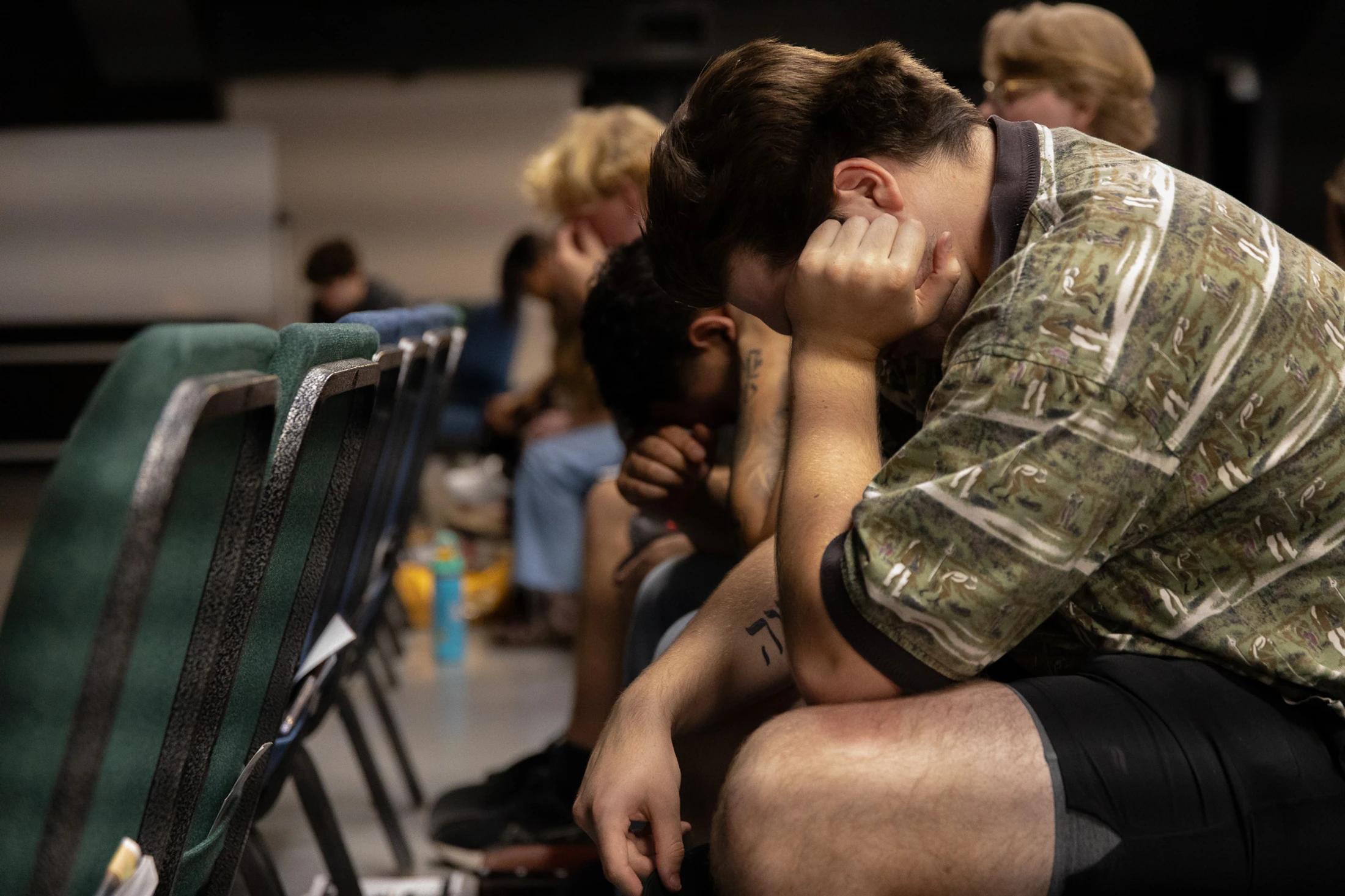 A side view of a young man in a patterned shirt sitting in a chair, bowing his head into his hands in prayer or deep thought, with others blurred behind him.