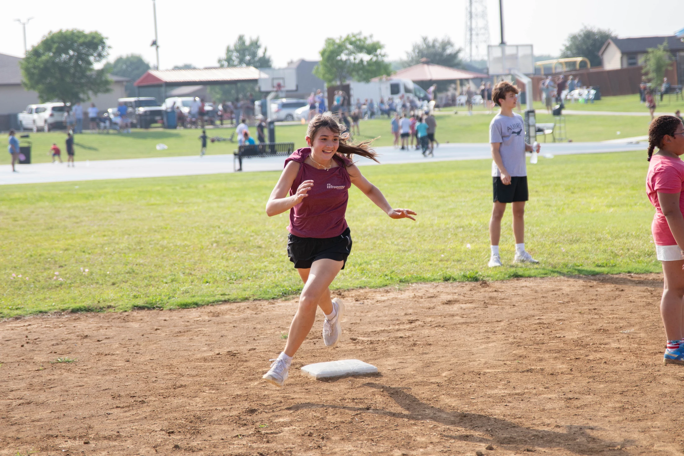 A smiling young girl in a maroon shirt and black shorts runs on a dirt baseball path towards a base during a sunny day, with others in the background.