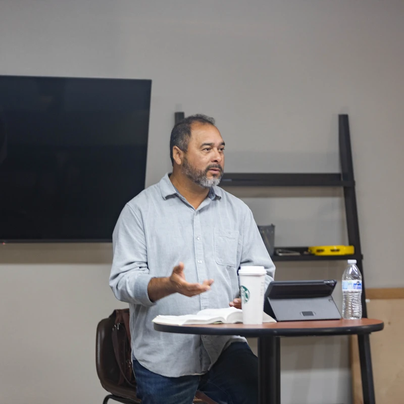 A man with a graying beard stands behind a table speaking, gesturing with his hand. On the table is an open book, a coffee cup, and a tablet.
