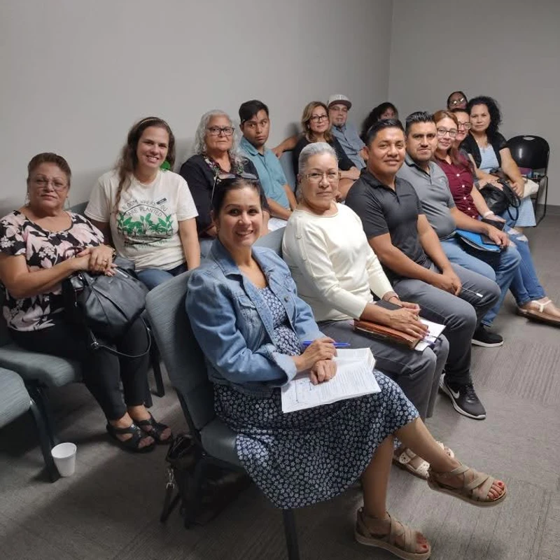 A diverse group of adults sit in several rows of chairs inside a room with plain walls, attentively looking towards the camera as if posing for a photo.