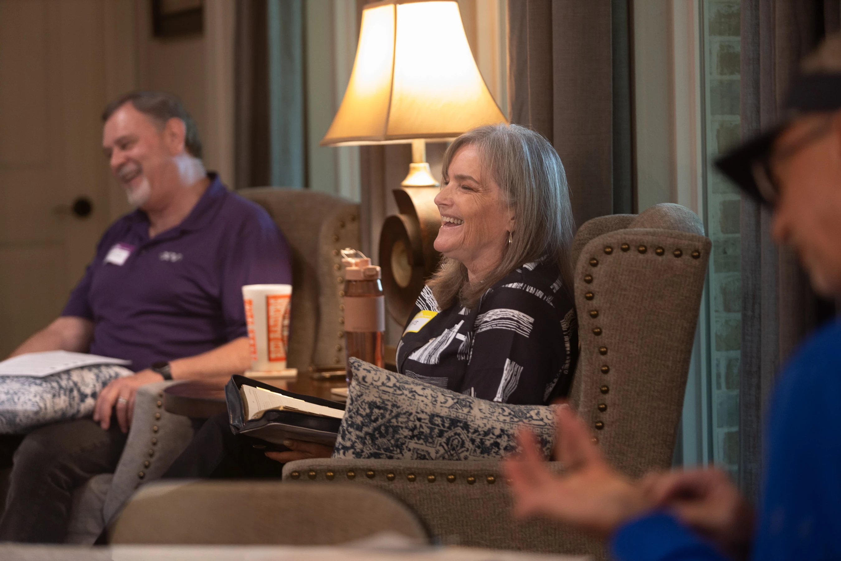 A gray-haired woman laughs while sitting in an armchair during a meeting in a warmly lit room. A man in the background also smiles.
