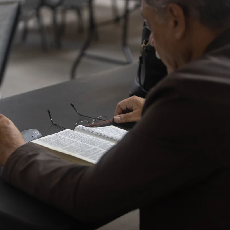 An over-the-shoulder view of a man in a brown jacket sitting at a table and reading an open Bible. He holds his glasses over the pages as he studies.