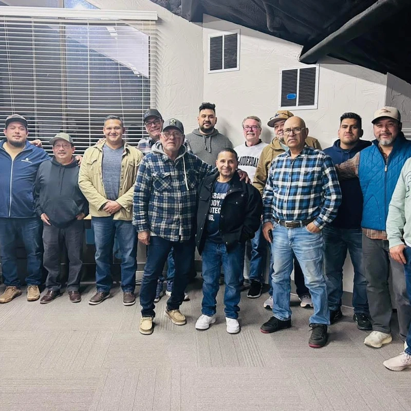 A large, diverse group of men of various ages standing together in two rows, smiling for a picture in a brightly lit room with a carpeted floor.