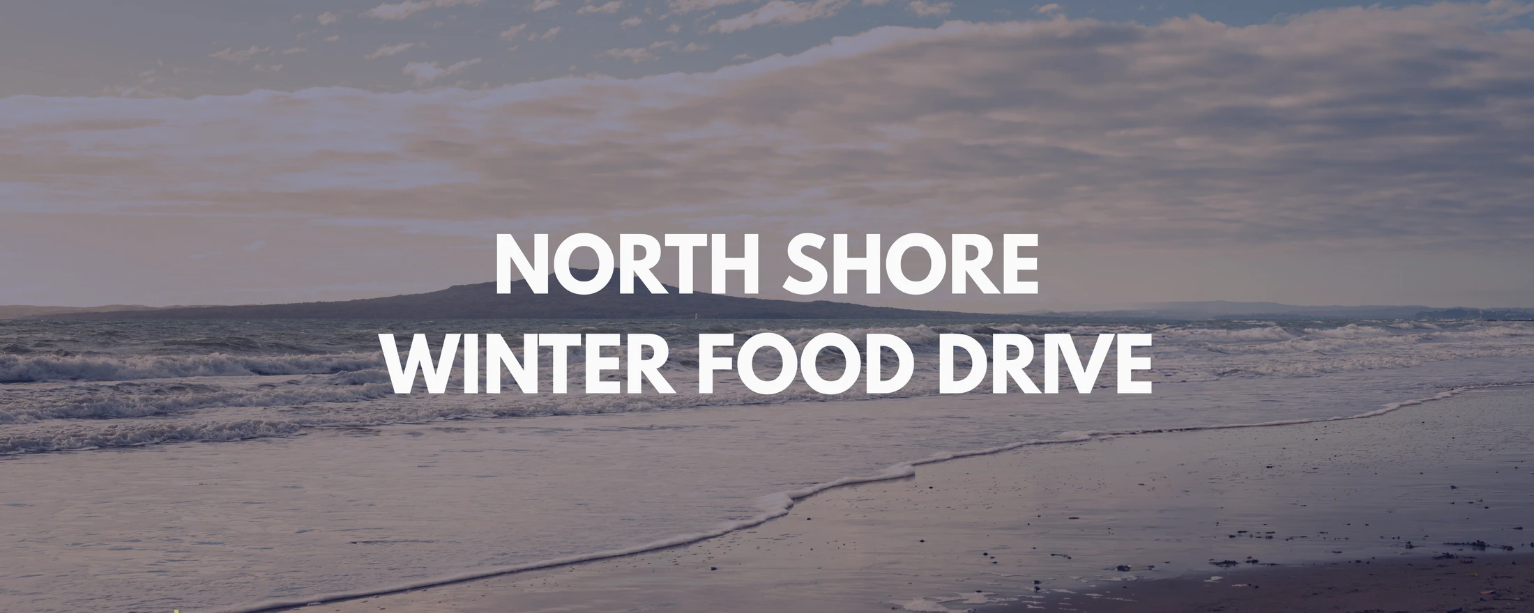 An image of a beach with waves rolling ashore under a cloudy sky. White text reads 'NORTH SHORE WINTER FOOD DRIVE' over the center of the image.