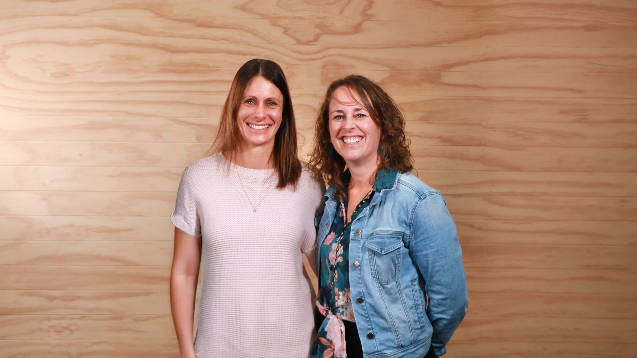 A medium shot of two smiling women standing side-by-side. One wears a pink sweater, and the other wears a denim jacket over a floral shirt, against a wood wall.