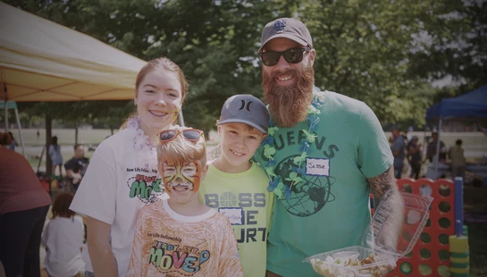 A smiling father with a beard and sunglasses poses with his three children at an outdoor event. One young boy has his face painted like a tiger.