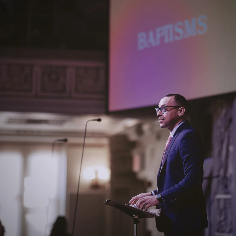 A man in a suit and glasses stands at a lectern speaking, with a screen behind him that reads 'BAPTISMS' in a dimly lit hall.