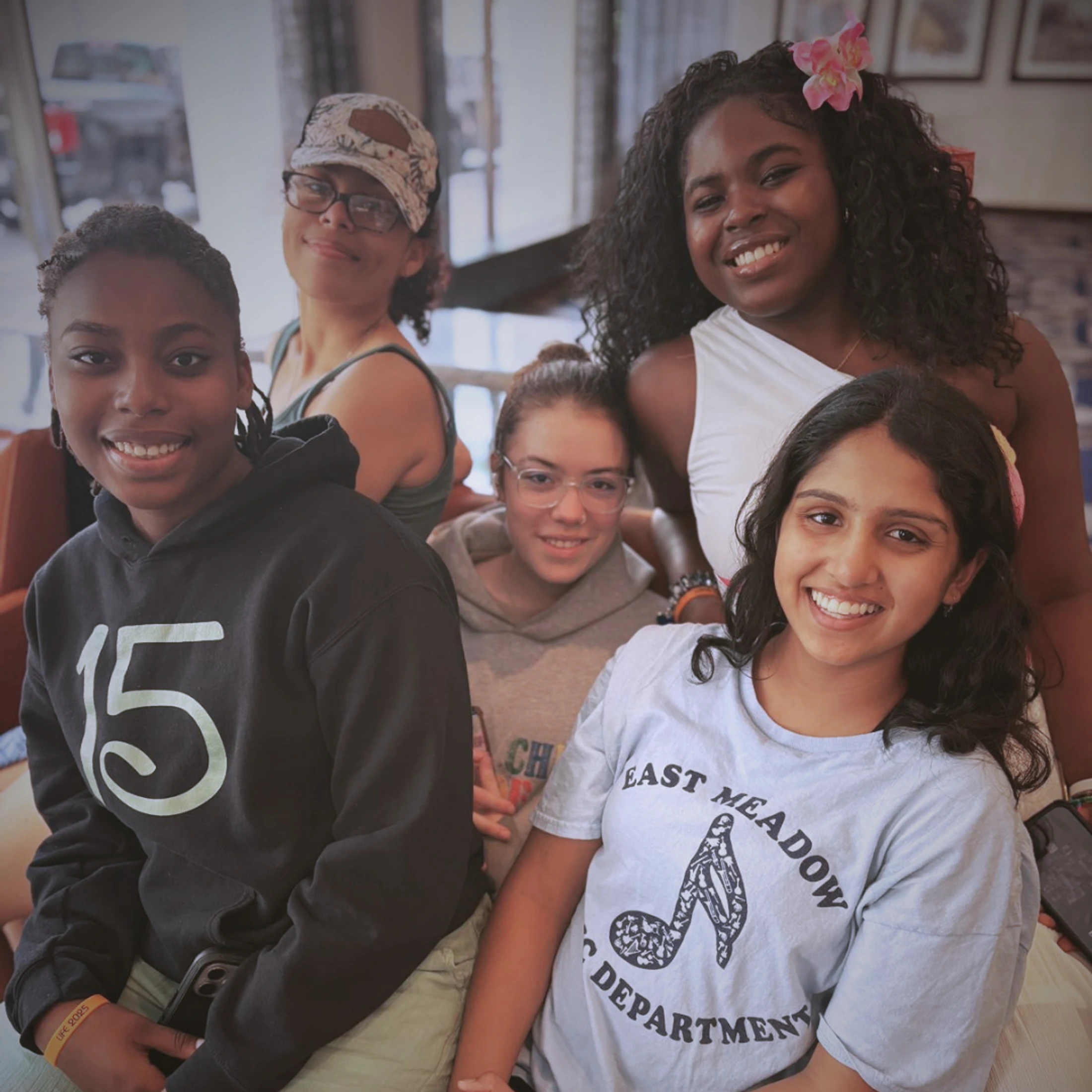 A diverse group of five smiling young women pose closely together for a photo indoors. They are all looking at the camera with happy expressions.