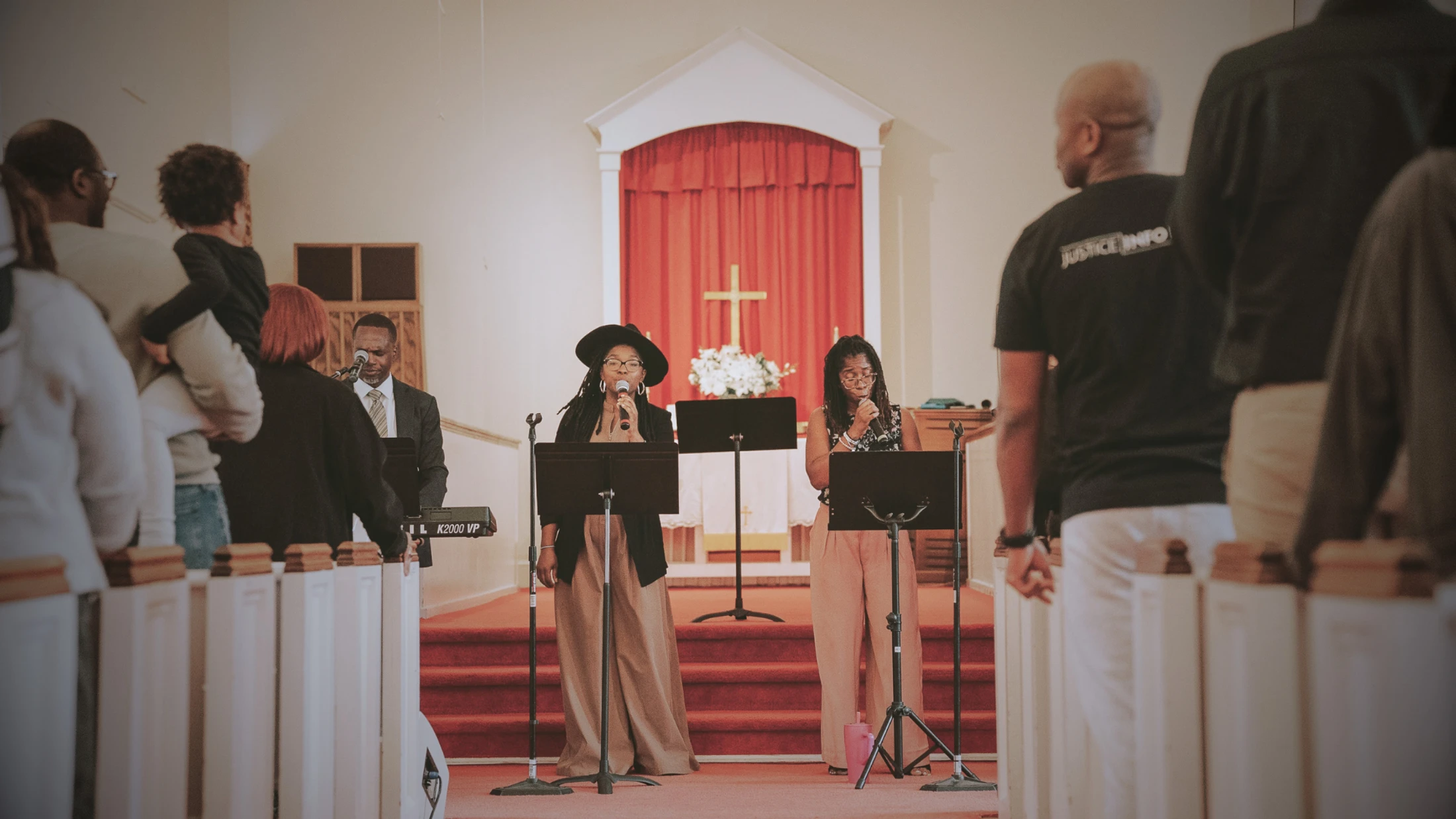 A view from the aisle of a church service where two women lead worship songs at the altar. A man plays keyboard and the congregation stands and listens.
