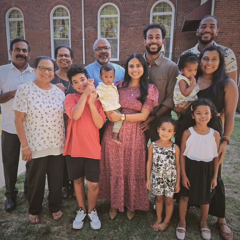 A large, smiling multi-generational family poses for a group photo on a lawn in front of a red brick building with arched windows.