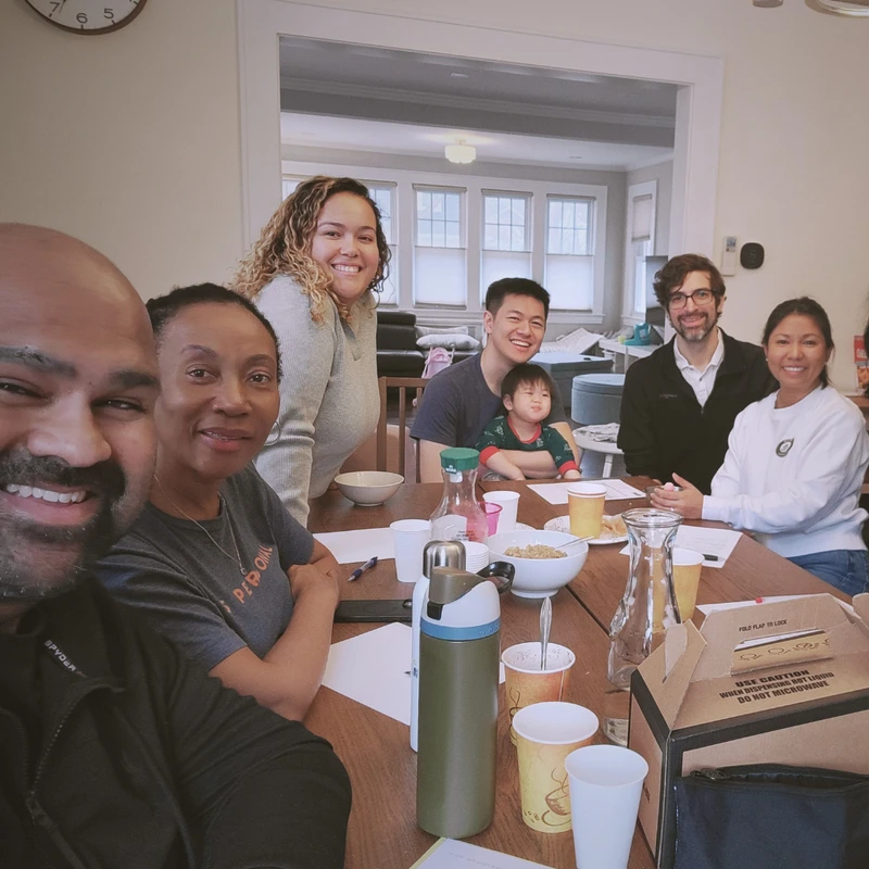 A smiling, diverse group of seven adults and one child posing for a selfie around a wooden dining table in a bright, cozy room.