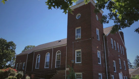 A low-angle view of a large, red brick church with a square tower against a bright blue sky. Green tree leaves frame the top of the sunny scene.