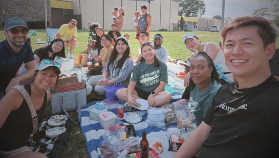 A large, diverse group of adults and children smiling for a selfie while sitting on blankets and enjoying a picnic on a grassy lawn on a sunny day.