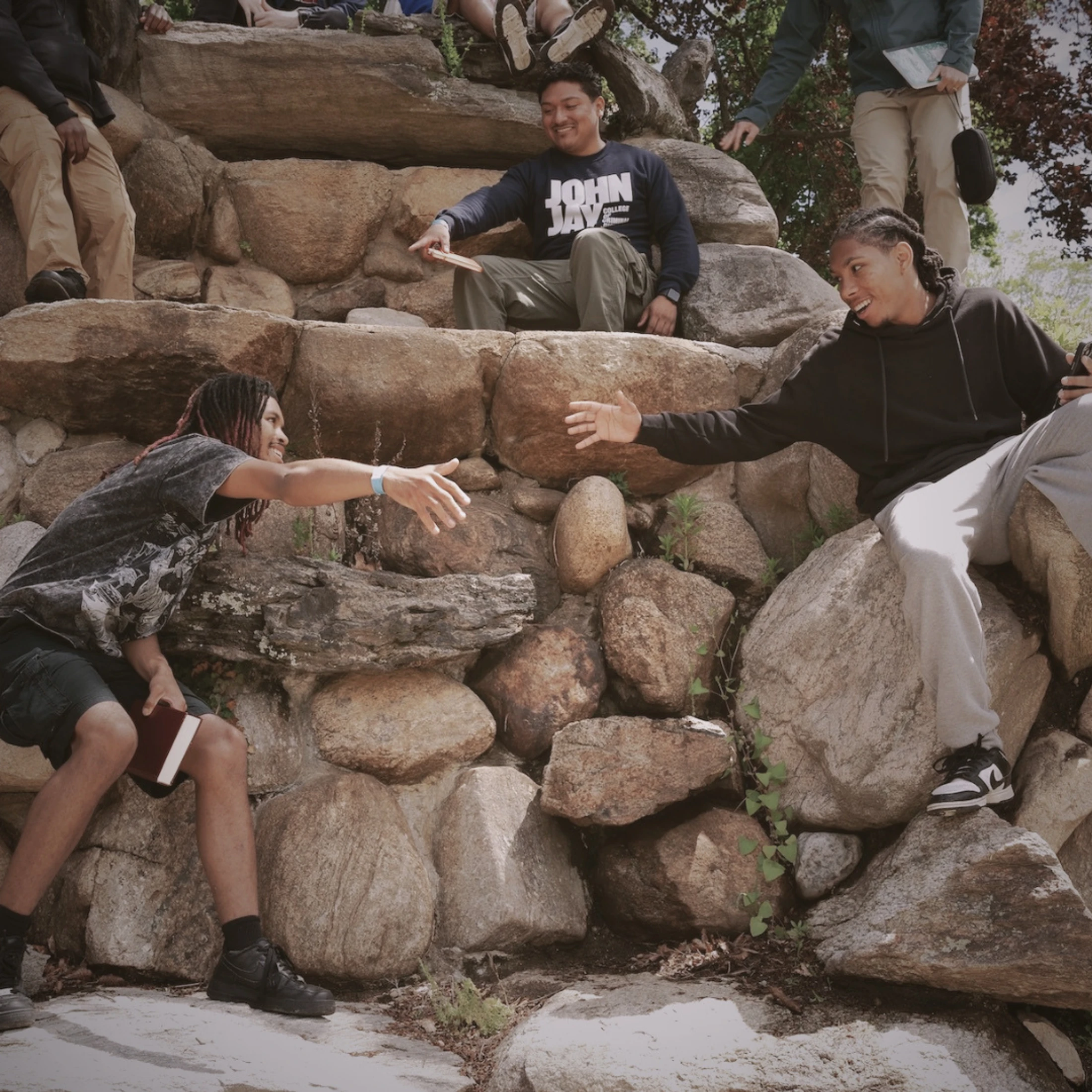 A group of young men are sitting on a large rock wall outdoors. Two men smile as they reach their hands out to one another in a gesture of friendship.