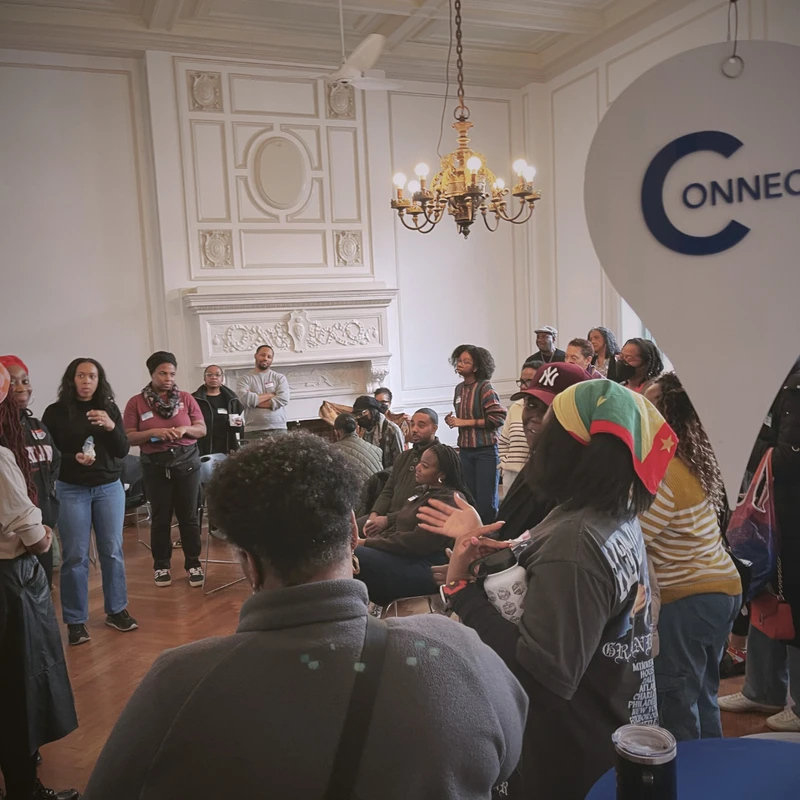 A diverse group of Black people stand and sit in a circle in an elegant room, engaged in a conversation during a community event.