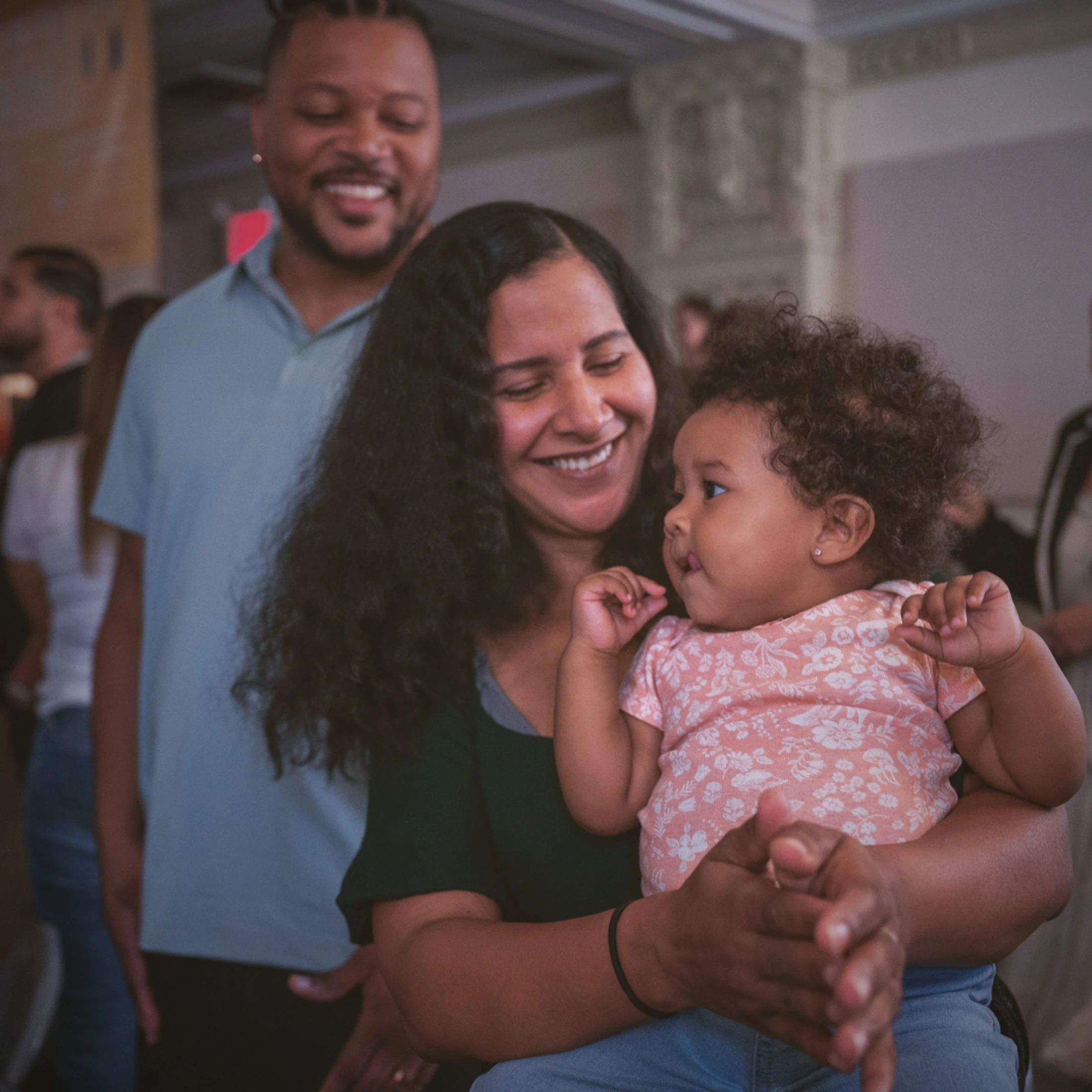 A smiling woman holds a baby girl who looks up at her. A smiling man stands behind them, creating a warm, happy family portrait.