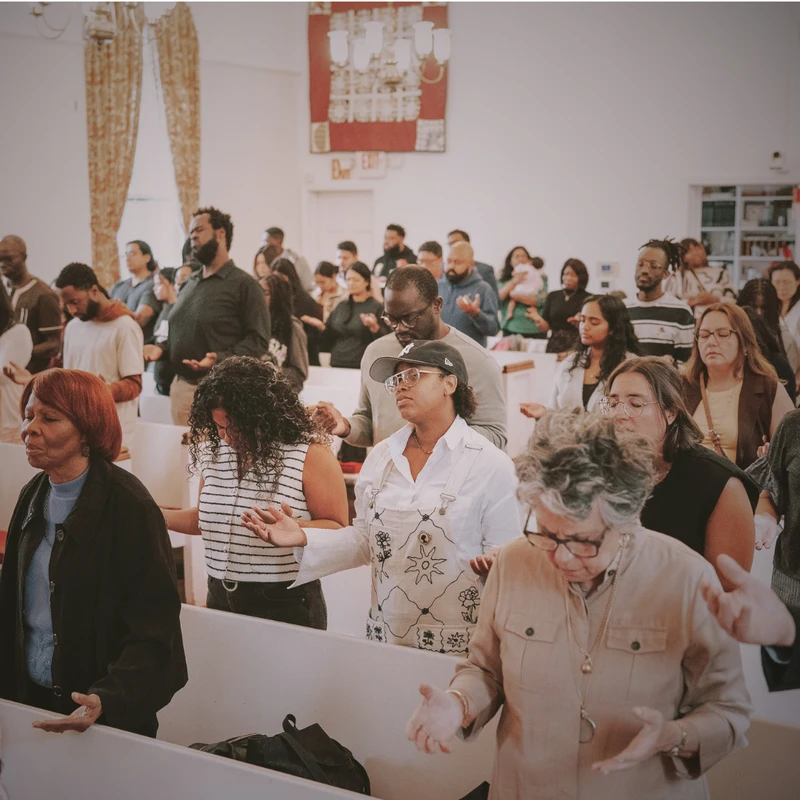 A diverse group of people stand in white pews inside a church, many with their eyes closed and hands raised in prayer and worship.