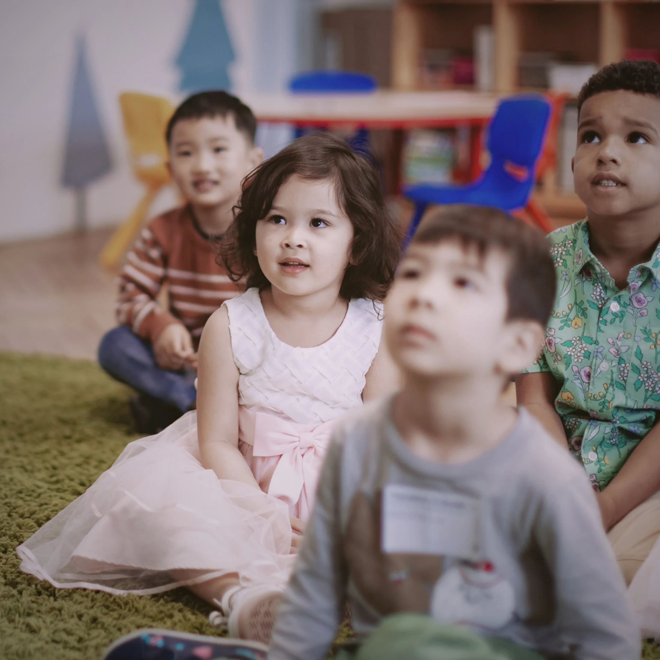 A diverse group of young children sit on a green carpet in a classroom, looking up with attentive expressions, with a girl in a pink dress in focus.