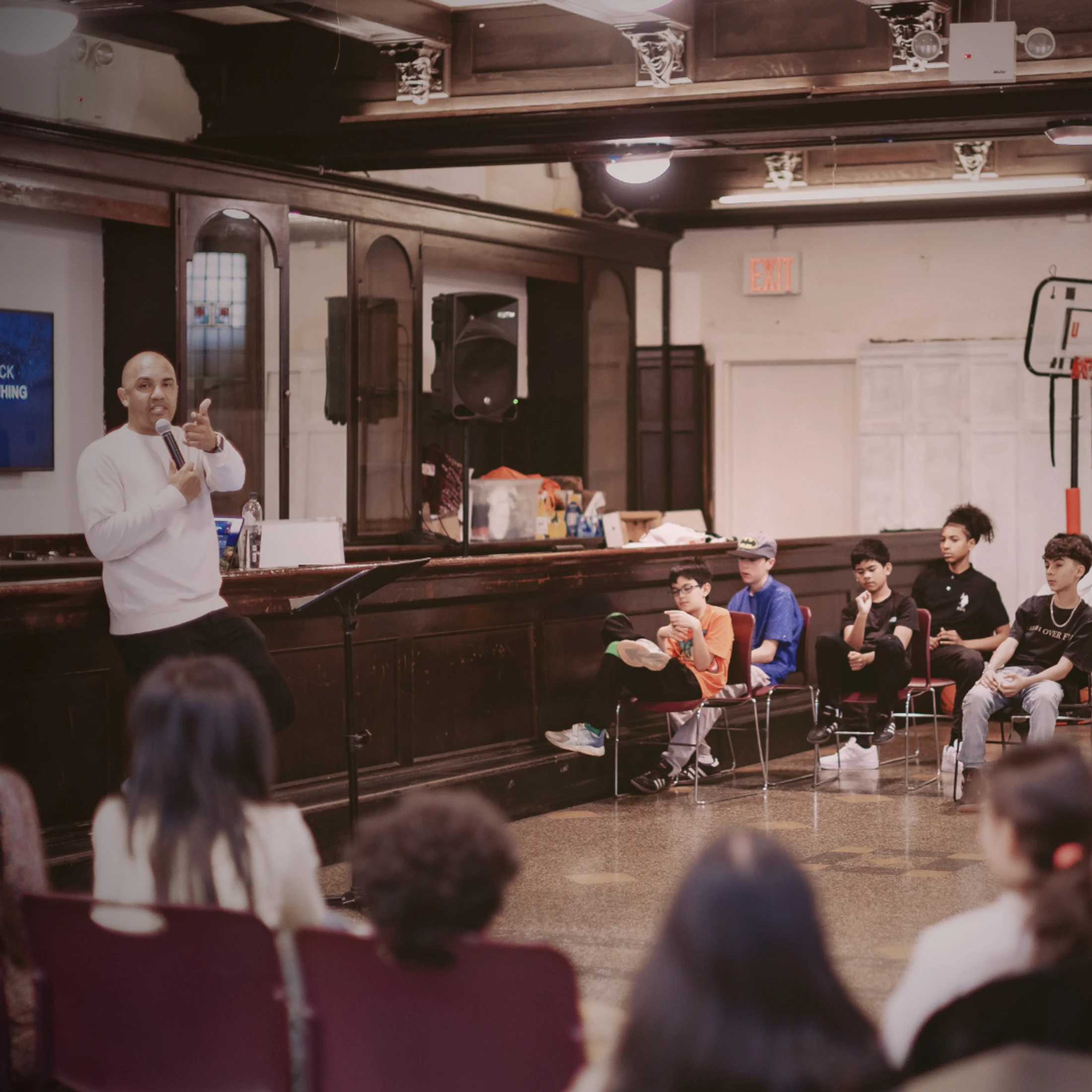 A man in a white sweatshirt speaks into a microphone, giving a thumbs-up to an audience of young people sitting in a large room with wood paneling.