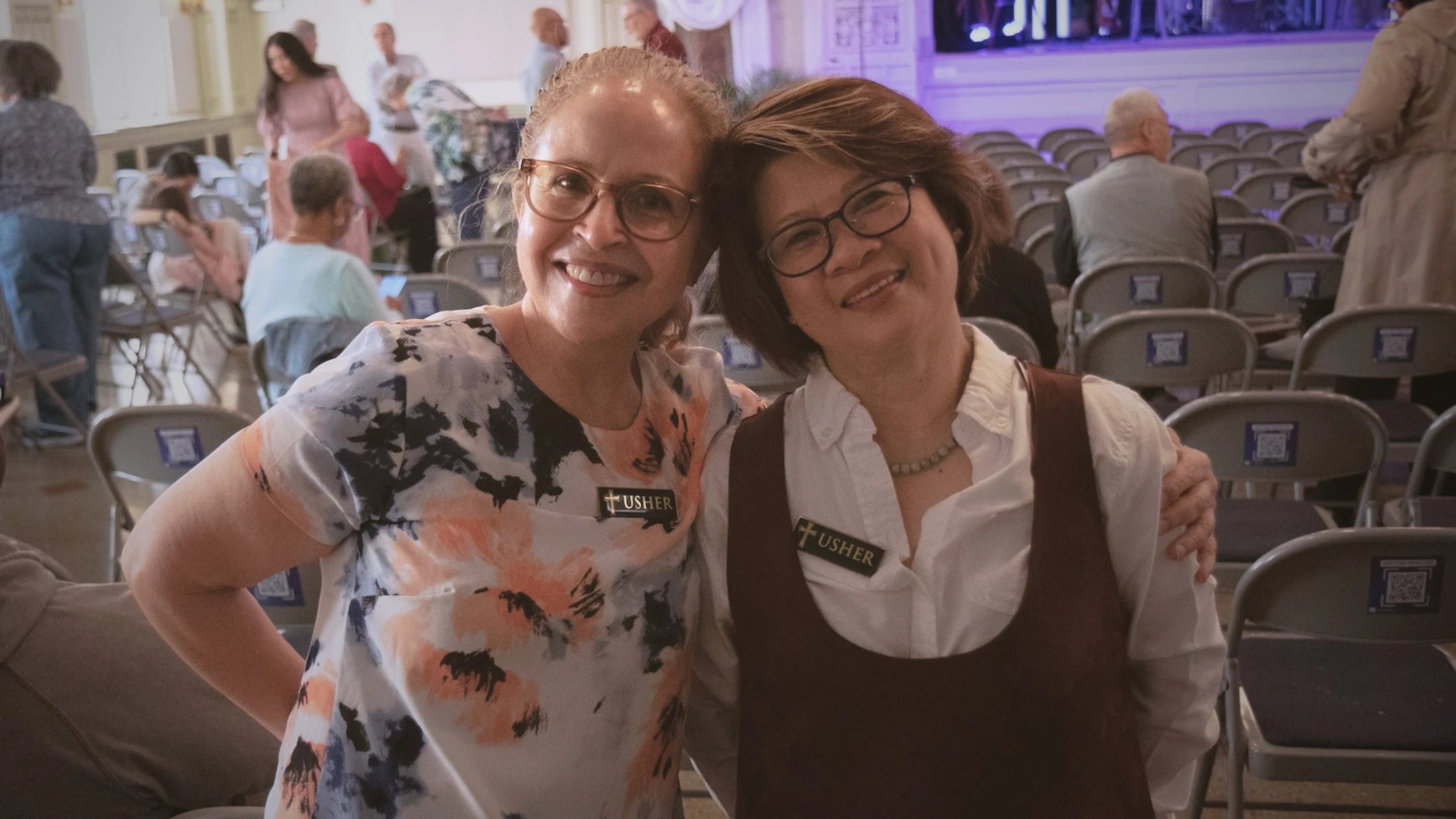 Two smiling female ushers with arms around each other pose for a photo in an auditorium filled with chairs and people before an event.
