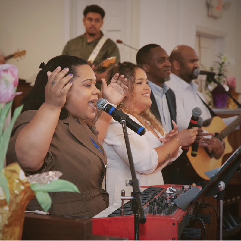 A woman in a brown blazer passionately sings into a microphone, leading a diverse worship band with other singers, guitarists, and a keyboard player.