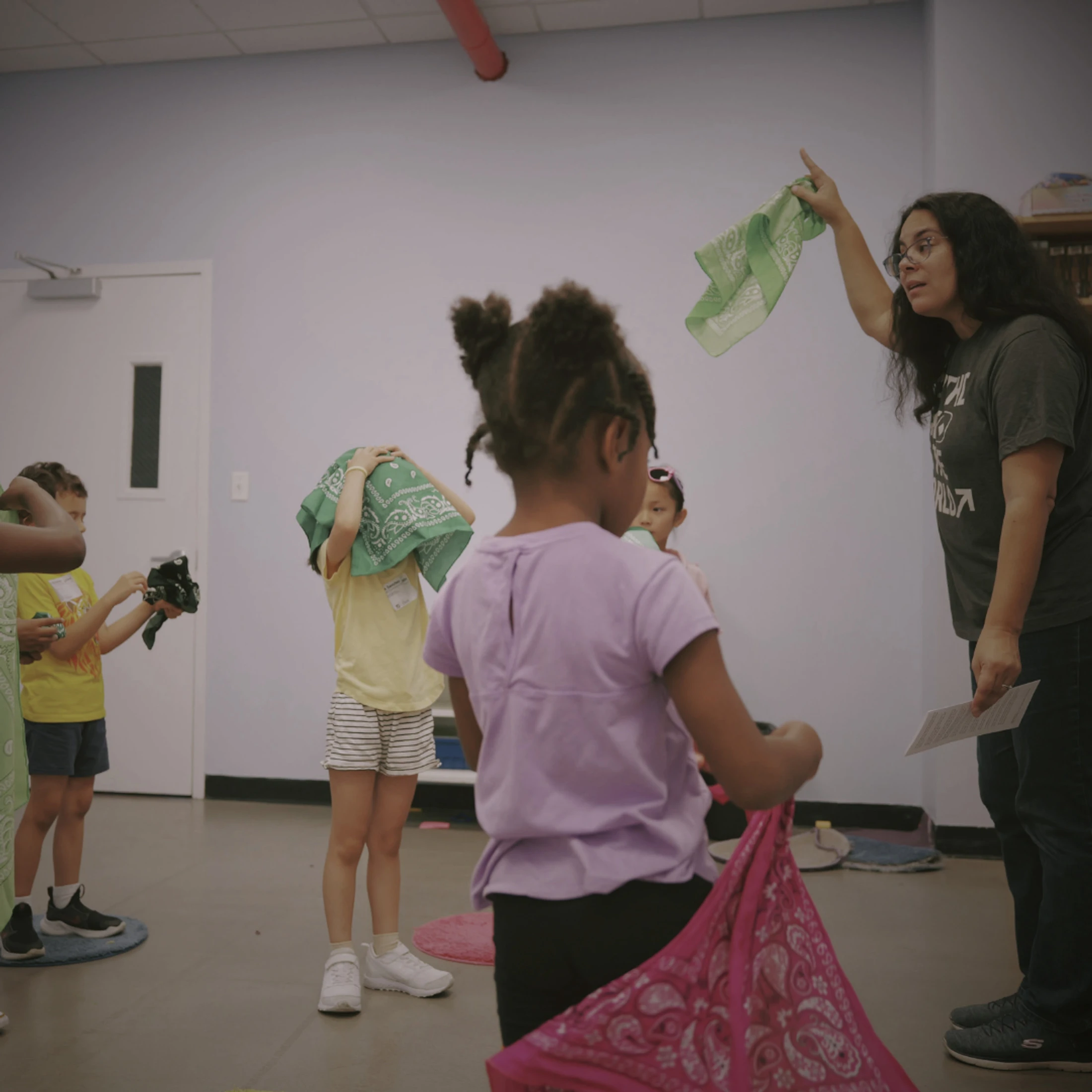 A woman instructs a group of diverse young children in a classroom setting. She holds up a green bandana, and the children interact with their own bandanas.