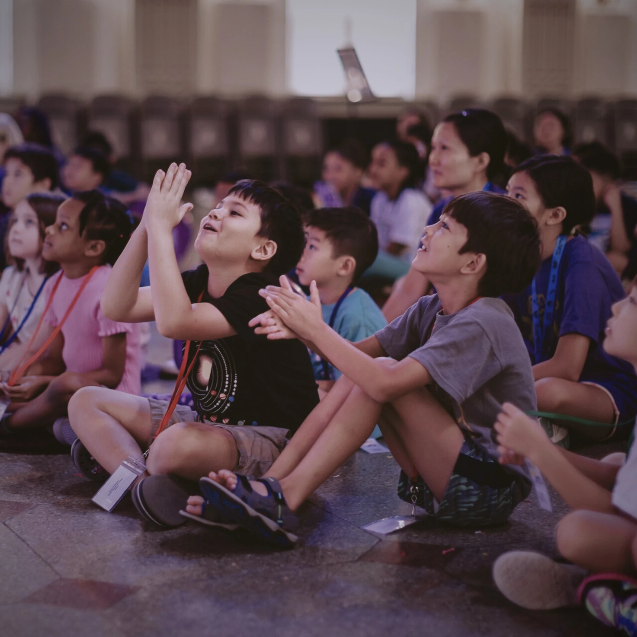 A diverse group of children sit on the floor in an auditorium, looking up with captivated expressions of wonder and excitement at something off-camera.