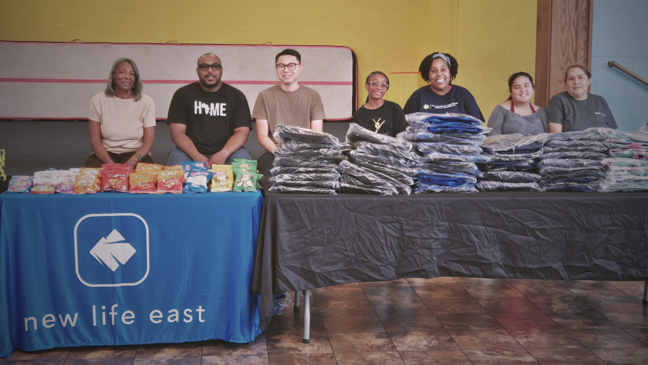 A diverse group of seven smiling volunteers sits behind tables loaded with snacks and packaged clothing at a 'new life east' community outreach event.