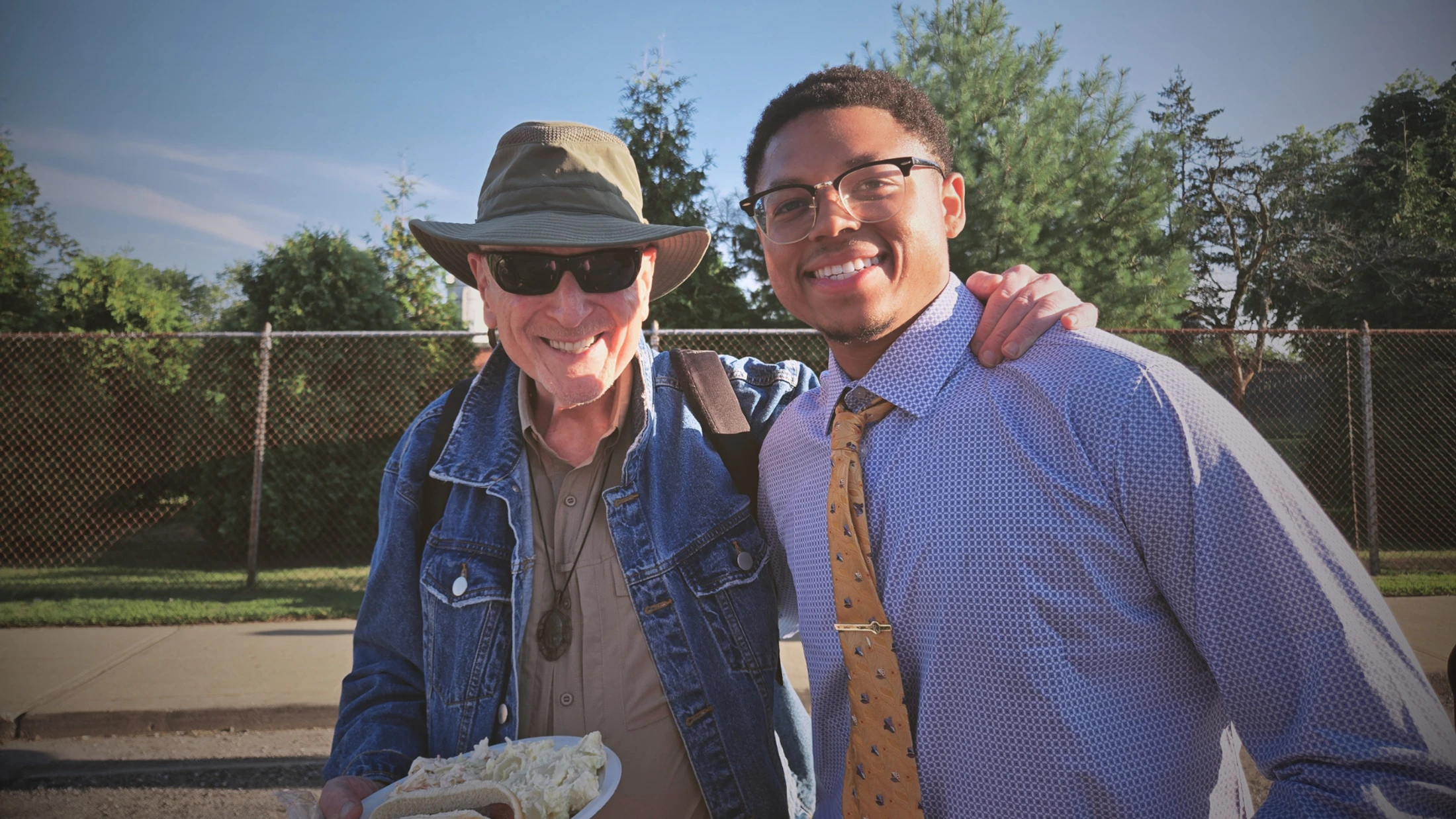 An older man in a hat and a younger man in a tie smile for the camera with their arms around each other outdoors, in front of a chain-link fence.