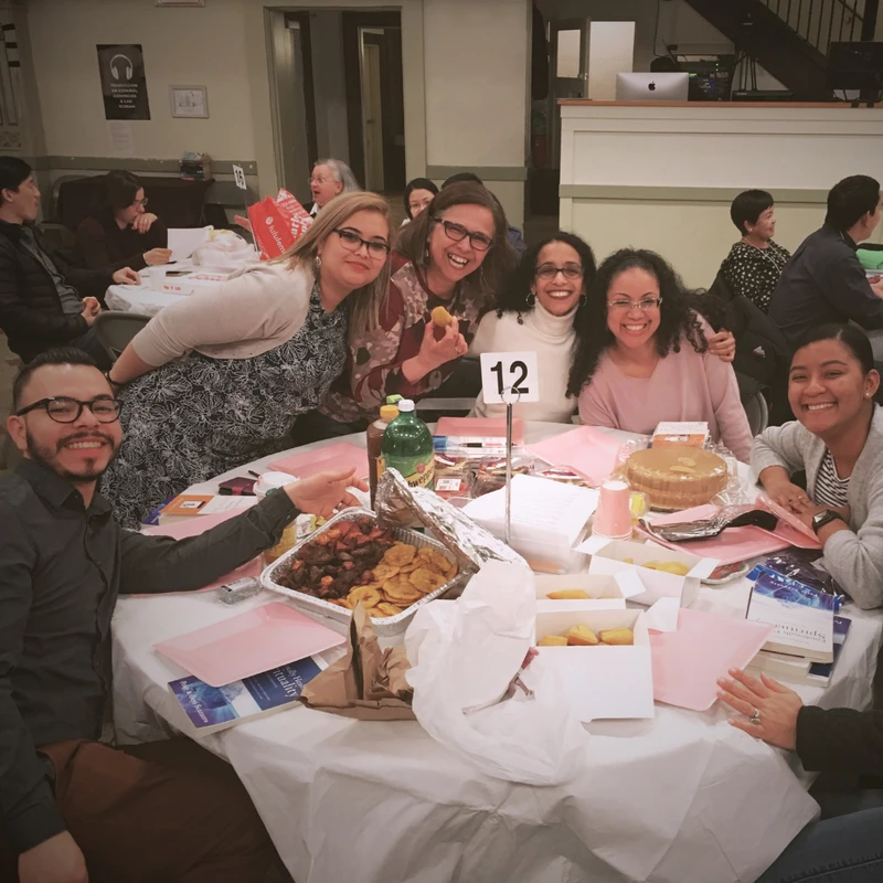 A diverse group of seven smiling people pose for a photo around a round table filled with food at a community event. The table has a number '12' sign.