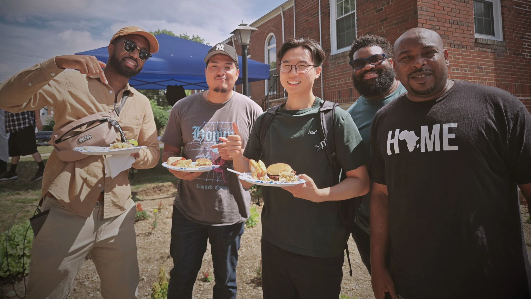 A diverse group of five men stand outside in front of a brick building, smiling at the camera while holding paper plates of food at a sunny cookout.