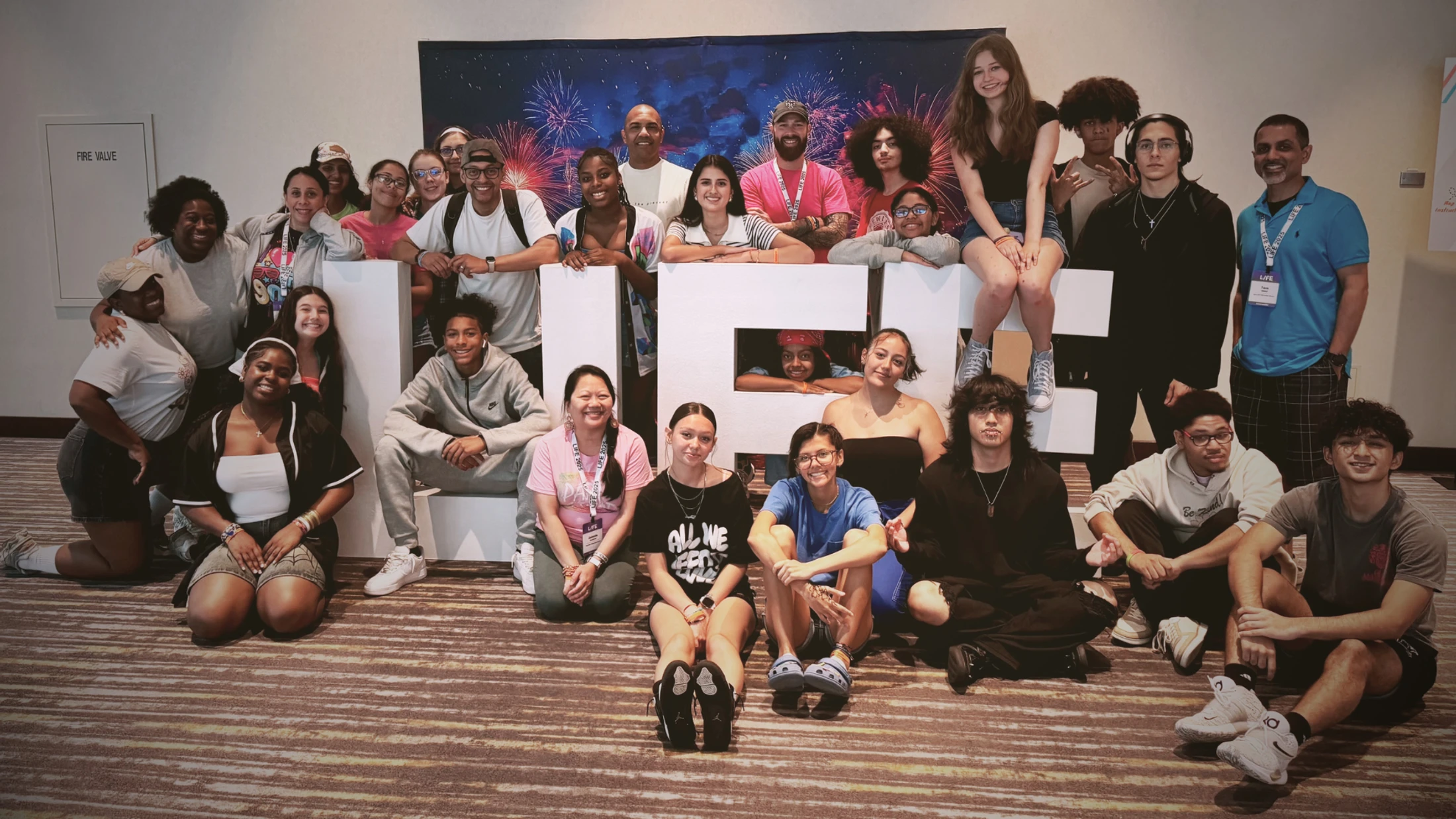 A large, diverse group of smiling teenagers and adults pose for a photo around large white letters spelling 'LIFE' at an indoor conference.