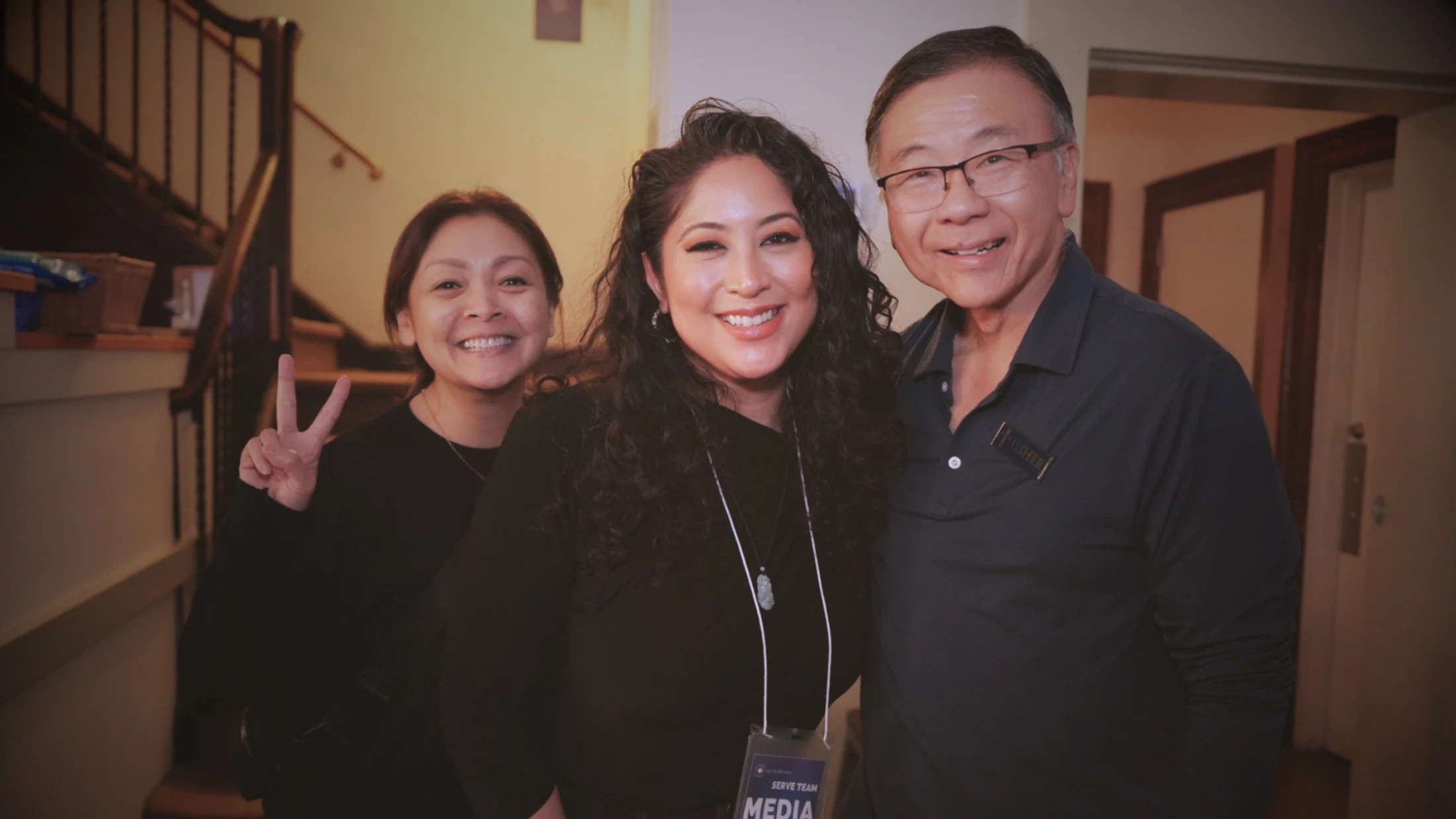 A warm, indoor photo of two women and a man smiling at the camera. The woman on the left makes a peace sign, and the woman in the middle wears a 'Media' badge.