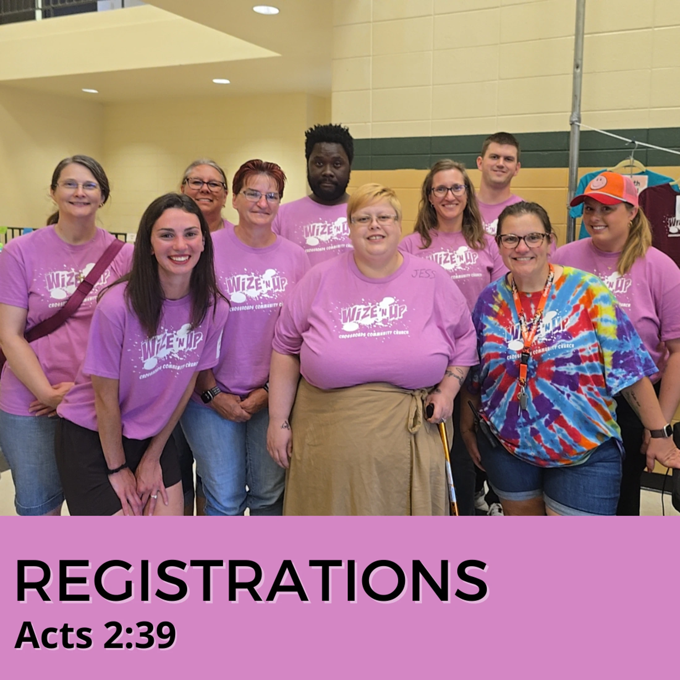 A group of smiling volunteers wearing matching purple 'Wize'n Up' t-shirts pose for a photo at a registration event in a community hall.