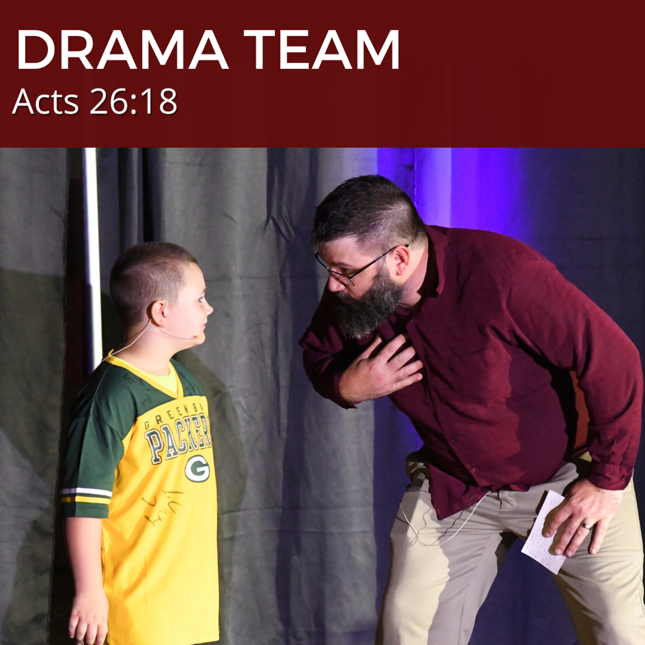 A man with a beard and a young boy act on a stage. The man leans in, gesturing to his chest, while the boy in a Packers jersey looks at him.